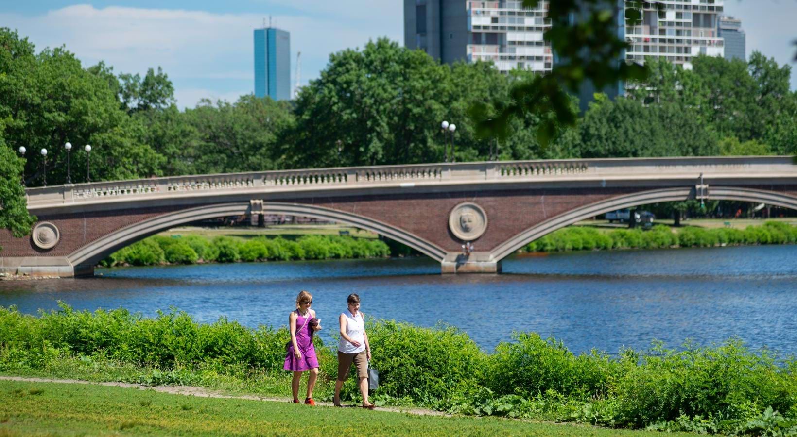 two women walking on a trail with a bridge in the background